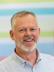 Dr Steven Wallis Smiling grey bearded man in front of pastel blue, green and pink horizontal striped background.