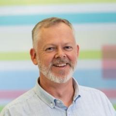 Smiling grey bearded man in front of pastel blue, green and pink horizontal striped background.